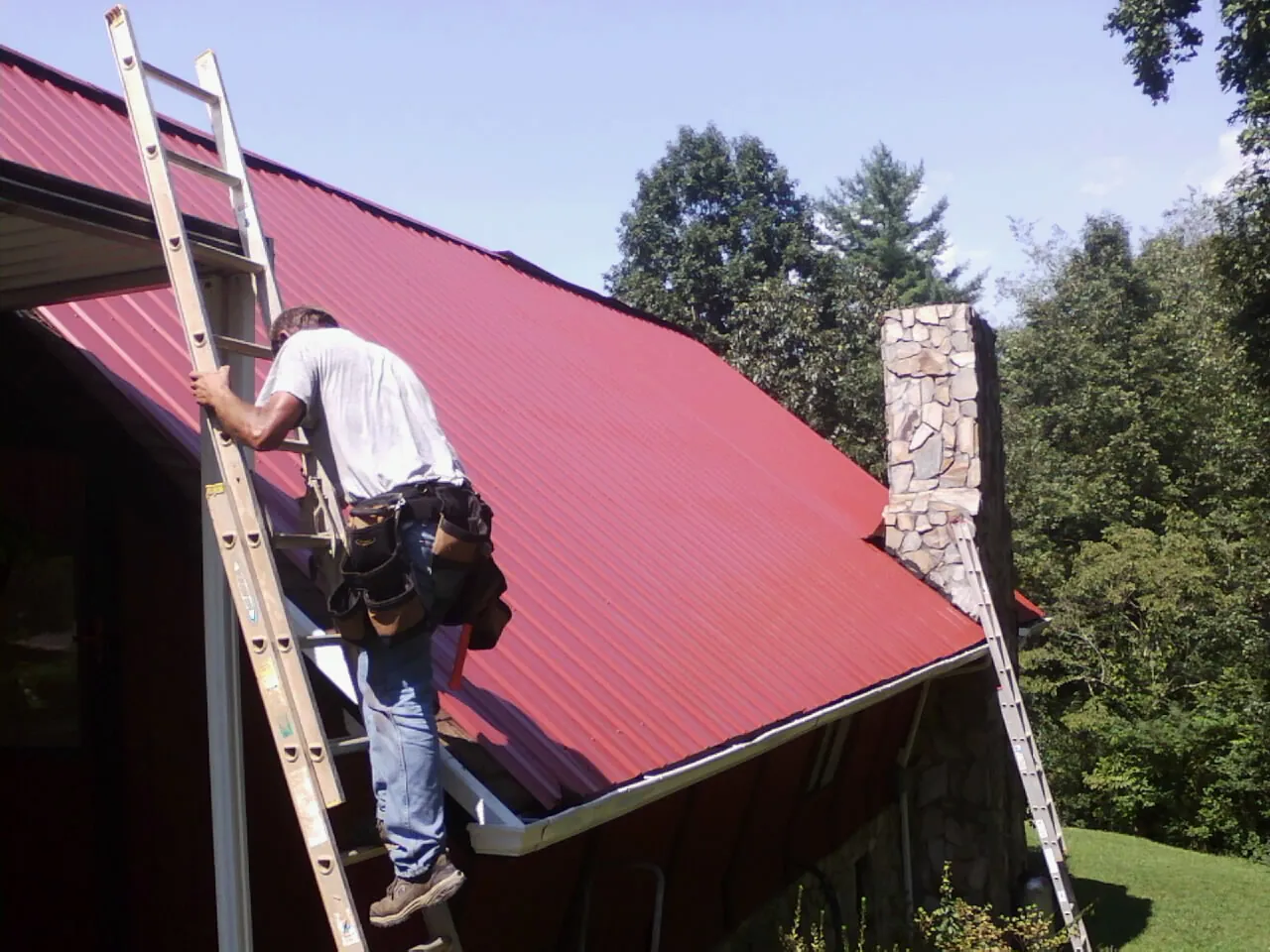Roofer on a ladder beside a red metal roof and stone chimney in Cummington